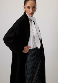Lungo cardigan nero a maglia sopra una camicia bianca con dettaglio del papillon, abbinato a una gonna di pelle nera con righe verticali.