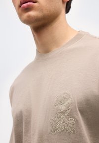 Κοντινό πλάνο ενός ατόμου που φοράει μπεζ πουκάμισο με κεντητό λουλούδι και στυλιζαρισμένο λογότυπο "Levi's" στ ο στήθος.