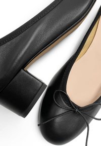 Ballerines en cuir noir avec un bout arrondi, un petit talon carré, une bordure élastique et un détail en nœud sur le devant. Texture lisse et design classique.