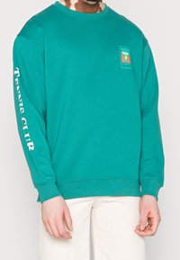 Sweatshirt bleu-vert en tissu doux avec un col rond, des manches longues, et un logo sur la poitrine ainsi que du texte le long de la manche.