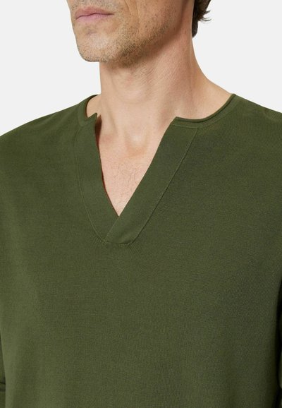 Hombre con suéter de punto verde oliva con escote en forma de muesca en V, mostrando la parte inferior del rostro y el cuello sobre un fondo liso.