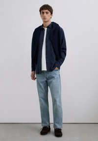 Camisa de botones navy sobre una camiseta blanca, combinada con jeans de un azul claro y zapatos de gamuza marrón. Diseño minimalist, tonos neutros.