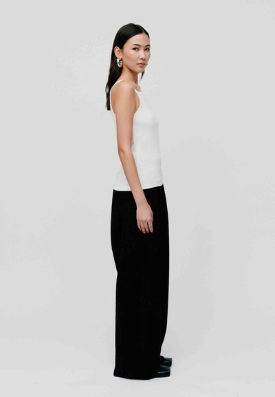 Top alb fără mâneci purtat cu pantaloni negri cu talie înaltă. Modelul stă de profil, design minimalist, textură netedă și siluetă simplă.