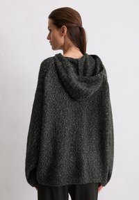 Pull oversized en tricot gris foncé avec une finition texturée et une capuche. Présente des épaules tombantes et des poignets côtelés. Silhouette décontractée.