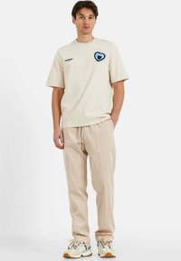 Camiseta de manga corta color beige con un gráfico de corazón en azul y verde, combinada con pantalones beige con cordón y zapatillas en tonos neutros.