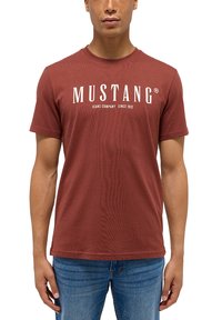 Červené bavlněné tričko s krátkými rukávy a kulatým výstřihem, s bílým nápisem "MUSTANG JEANS COMPANY SINCE 1932" na hrudi.