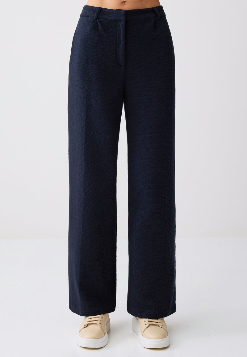 Pantalon large bleu marine porté avec des baskets beige à lacets, sur une personne debout devant un fond uni.
