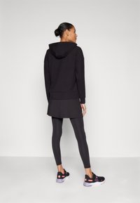 Sweatshirt à capuche noir, superposé sur une jupe noire, associé à des leggings noirs. Présente un matériau texturé et des accents de semelle contrastants sur les chaussures.