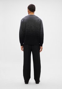 Homem em pé de costas, vestido com um suéter em gradiente preto e cinza, calças largas pretas e sapatos pretos, sobre um fundo branco.
