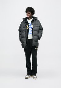 Czarna kurtka puchowa z kapturem, noszona na szarej bluzie z nadrukiem. Czarne spodnie dresowe z złotym logo "Nike" i szare trampki.