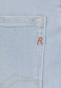 Tejido de mezclilla azul claro con un solo bolsillo trasero que presenta la letra "R" bordada en rojo, costuras visibles y una textura sutil.