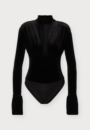 Femme aux cheveux noirs en queue de cheval basse portant un haut en velours noir à manches longues avec découpe et un pantalon noir à jambes larges, debout sur fond blanc.