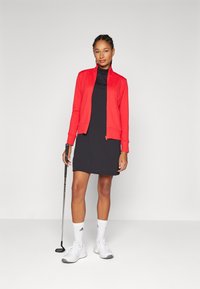 Jaqueta texturizada vermelha sobre um vestido preto, combinada com meias e sapatos brancos. Um taco de golfe é segurado com a mão direita.