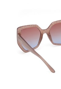 Lunettes de soleil en forme hexagonale avec une monture transparente teintée de rose et des verres dégradés passant du rose au bleu. Logo "GUESS" sur la branche.