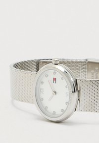 Zilveren polshorloge met mesh-band, witte wijzerplaat, kristallen uurmarkeringen, Romeinse cijfers op 6 en 12, en Tommy Hilfiger logo op de wijzerplaat.