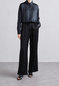 Svart silkeskjorta med stångknäppning och långa ärmar, kombinerad med lösa svarta satinbyxor och svarta sandaler med öppen tå och klack.