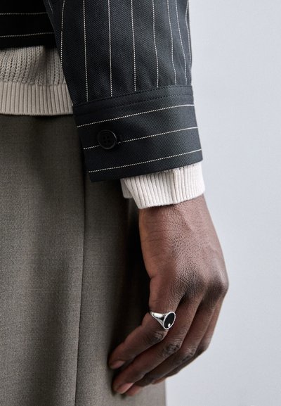 Polsino di giacca a righe nere con un bottone, sovrapposto a un maglione a maglia beige chiaro. Una mano che indossa un anello d'argento poggia su un tessuto grigio.