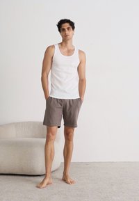 Weißes Tanktop und beiger Shorts auf einer stehenden männlichen Figur, barfuß, mit kurzem dunklem Haar, vor einem schlichten Hintergrund und einer weichen Couch.