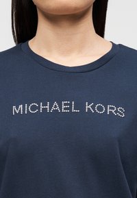 T-shirt de algodão azul-marinho com decote redondo e o texto "MICHAEL KORS" em tachas prateadas no peito. Textura lisa.