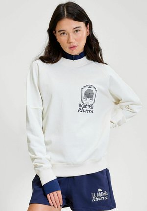 Młoda kobieta o długich ciemnych włosach, ubrana w białą bluzę i granatowe szorty, oba z logo "Il Club Della Riviera".