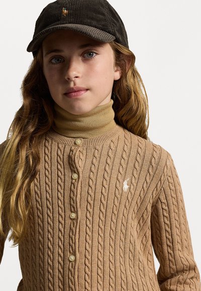 Κάπα με πλέξη κασμίρ σε καφέ απόχρωση με κουμπί στο μπροστινό μέρος και μικρό λευκό λογότυπο. Συνδυάζεται με μπεζ ζιβάγκο, φορεμένο κάτω από σκουρόχρωμο καπέλο από κορντούρα.