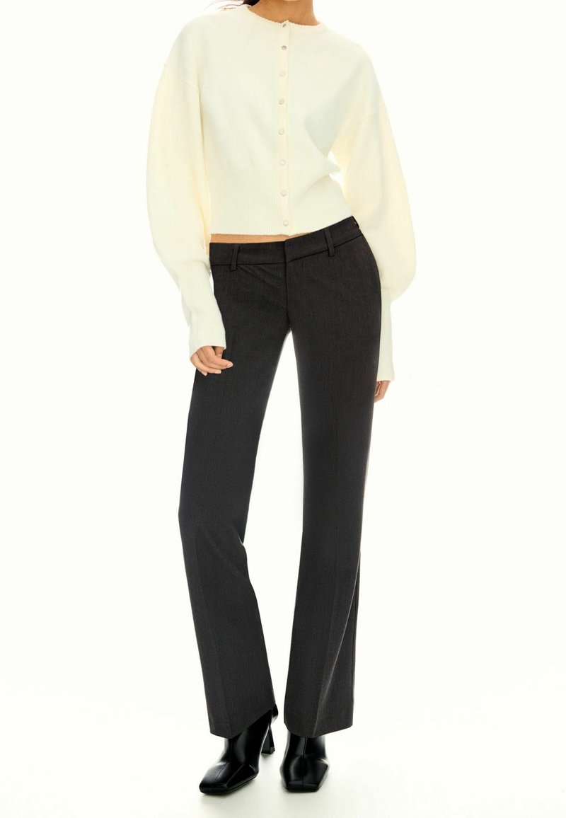 Femme portant un cardigan blanc boutonné avec des manches ballon, un pantalon droit foncé et des bottines noires à talons.