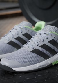 Paire de chaussures de sport Adidas grises avec des bandes noires et une doublure intérieure verte, placées sur une surface noire texturée près d'une plaque de poids.