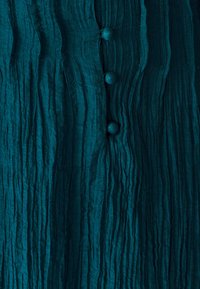 Teal-farbenes, strukturiertes Stoffblusengewand mit vertikalen Falten und vier passenden, runden Knöpfen auf der Vorderseite, die ein zerknittertes Aussehen zeigen.