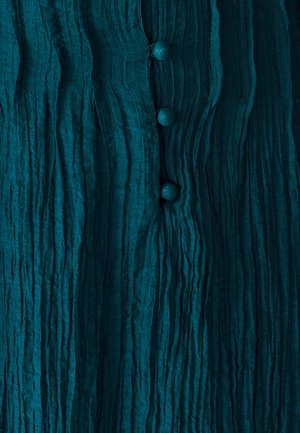 Teal textuurstof blouse met verticale plooien en vier bijpassende ronde knopen aan de voorkant, met een gekreukt uiterlijk.