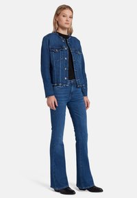 Giacca di jeans blu scuro con orli sfilacciati, bottoni argentati e tasche sul petto, abbinata a jeans blu a gamba svasata e stivaletti neri alla caviglia.