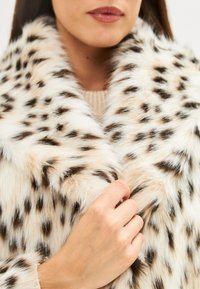 Műszőr kabát, krémszínű alappal, fekete és barna leopárd pöttyökkel, széles gallérral és puha textúrával. Kéz a gallért fogja.