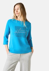 Helles blaues Top mit drei Viertelärmeln, das ein geometrisches schwarzes Grafikdesign und Text zeigt, kombiniert mit grauen Sweatpants aus weichem Stoff.