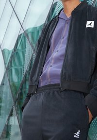 Chaqueta bomber negra de textura suave, combinada con una camisa morada abotonada con rayas verticales y pantalones deportivos negros a juego.