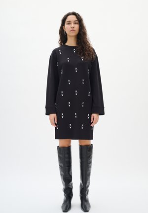 Schwarzes Sweatshirtkleid mit silbernen Verzierungen, langen Ärmeln, Rundhalsausschnitt und knielanger Länge. Getragen mit hohen schwarzen Lederstiefeln.