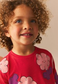 Tricou roșu cu exotic, cu desene mari de flori zâmbitoare în roz și violet. Material moale, cu guler rotund și mâneci scurte.