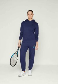 Felpa con cappuccio blu navy e pantaloni sportivi realizzati in tessuto morbido, con un logo sul petto e un dettaglio coordinato sulla gamba. Tiene una racchetta da tennis.