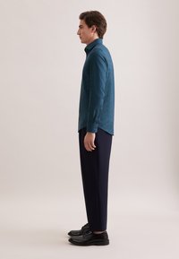 Turkooise overhemd met knopen, een gestructureerd patroon, lange mouwen en zwarte nette broek. Model staat zijwaarts, waardoor de pasvorm en kraag van het overhemd goed zichtbaar zijn.