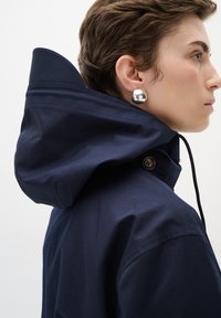 Jūras zilas ūdensnecaurlaidīgas jakas ar augstu apkakli un plašu kapuci, ar podziņu akcentiem un regulējamu auklu, lai pielāgotu izmēru.