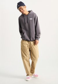 Sudadera gris con un bolsillo frontal y logo, combinada con pantalones beige holgados y zapatillas rosas. Modelo lleva un gorro oscuro.