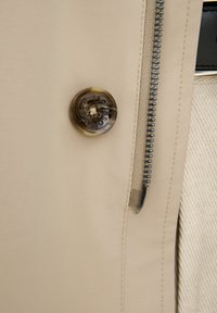 Tessuto beige con una cerniera visibile, accompagnato da un pulsante rotondo marmorizzato con dettagli intricati e una superficie liscia.