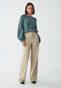 Ein türkisfarbener Strickpullover mit lockerer Passform, Ballonärmeln, kombiniert mit hochtaillierten beigen weitem Hosen und schwarzen spitzen Absatzschuhen.