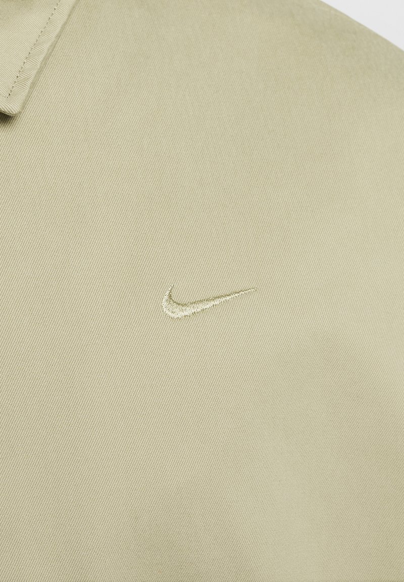 Tissu beige avec un logo swoosh Nike brodé, présentant un gros plan de la couture du col sur le coin supérieur gauche.