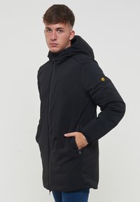 Giacca imbottita nera con cappuccio, due tasche laterali e logo accento sulla manica. Texture liscia e design di lunghezza media.