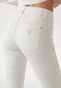 I jeans di denim bianco presentano un alto punto vita, una silhouette aderente e due tasche posteriori. Un piccolo logo triangolare è visibile su una tasca.