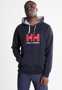 Sudadera azul marino con forro gris, cuenta con un bolsillo frontal y un gran logo de Helly Hansen en rojo y blanco, fabricada en material de algodón, corte relajado.