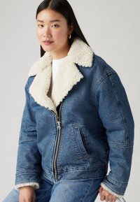 Chaqueta de mezclilla con cuello de borreguito crema, cremallera frontal, bolsillos laterales y un ajuste holgado. Textura de mezclilla azul con detalles de costura visibles.