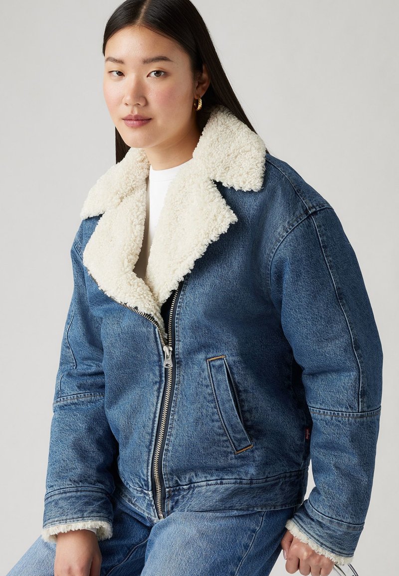 Veste en denim avec un col en shearling crème, fermeture éclair sur le devant, poches latérales et coupe décontractée. Texture en denim bleu avec des détails de couture visibles.