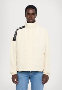 Polstret hvid jakke med høj krave, sort branding på skulderen, elastiske manchetter og en frontlynlås. Bæret med lyseblå jeans.