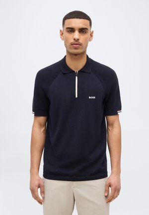 Άνδρας που φοράει πλεκτό πόλο με κοντά μανίκια σε ναυτικό μπλε χρώμα με μπροστινό φερμουάρ και λογότυπο "BOSS", συνδυασμένο με ανοιχτό μπεζ παντελόνι, στέκεται μπροστά σε λευκό φόντο.