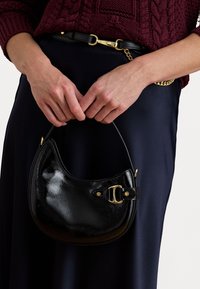 Fekete bőrtáska fényes felülettel, ívelt formával, arany hardware-rel és rövid fogantyúval, kézben tartva egy sötét ruházat mellett.